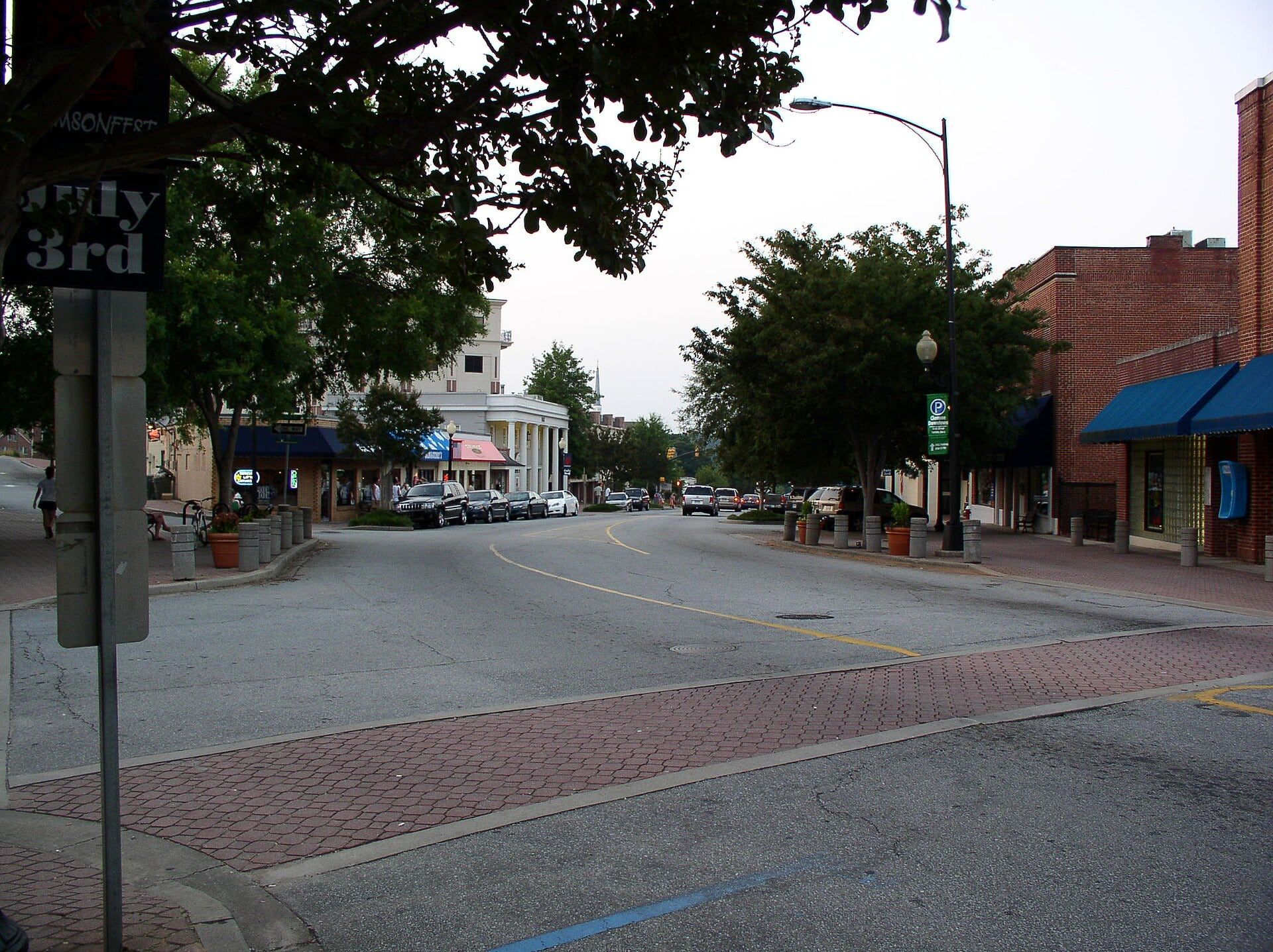 A street in downtown Clemson with brick shopfronts, blue awnings, parked cars and shade trees.